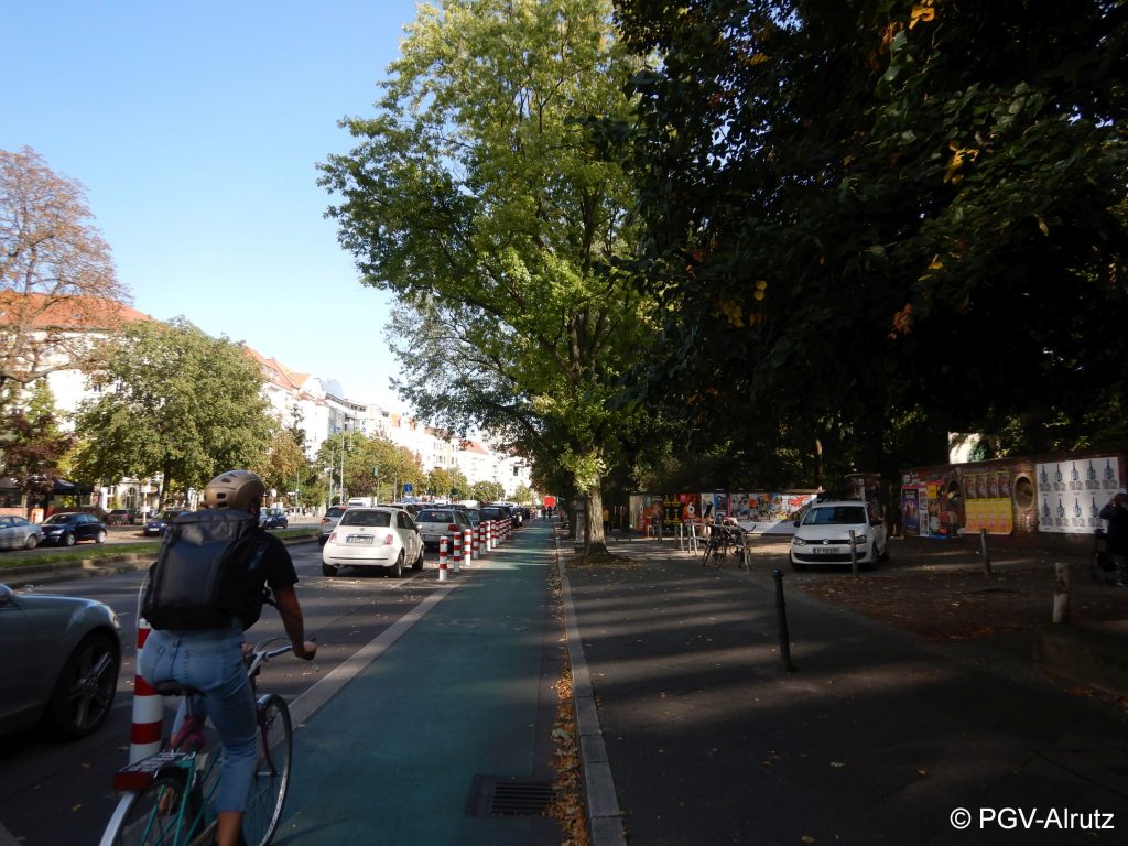 Das Bild zeigt einen geschützten Radfahrstreifen in Berlin. Links ist eine Reihe rot-weiß-gestreifte Poller, die den Radfahrstreifen vom Parkstreifen trennen. Auf dem Radfahrstreifen fährt eine Person Rad- Copyright PGV-Alrutz