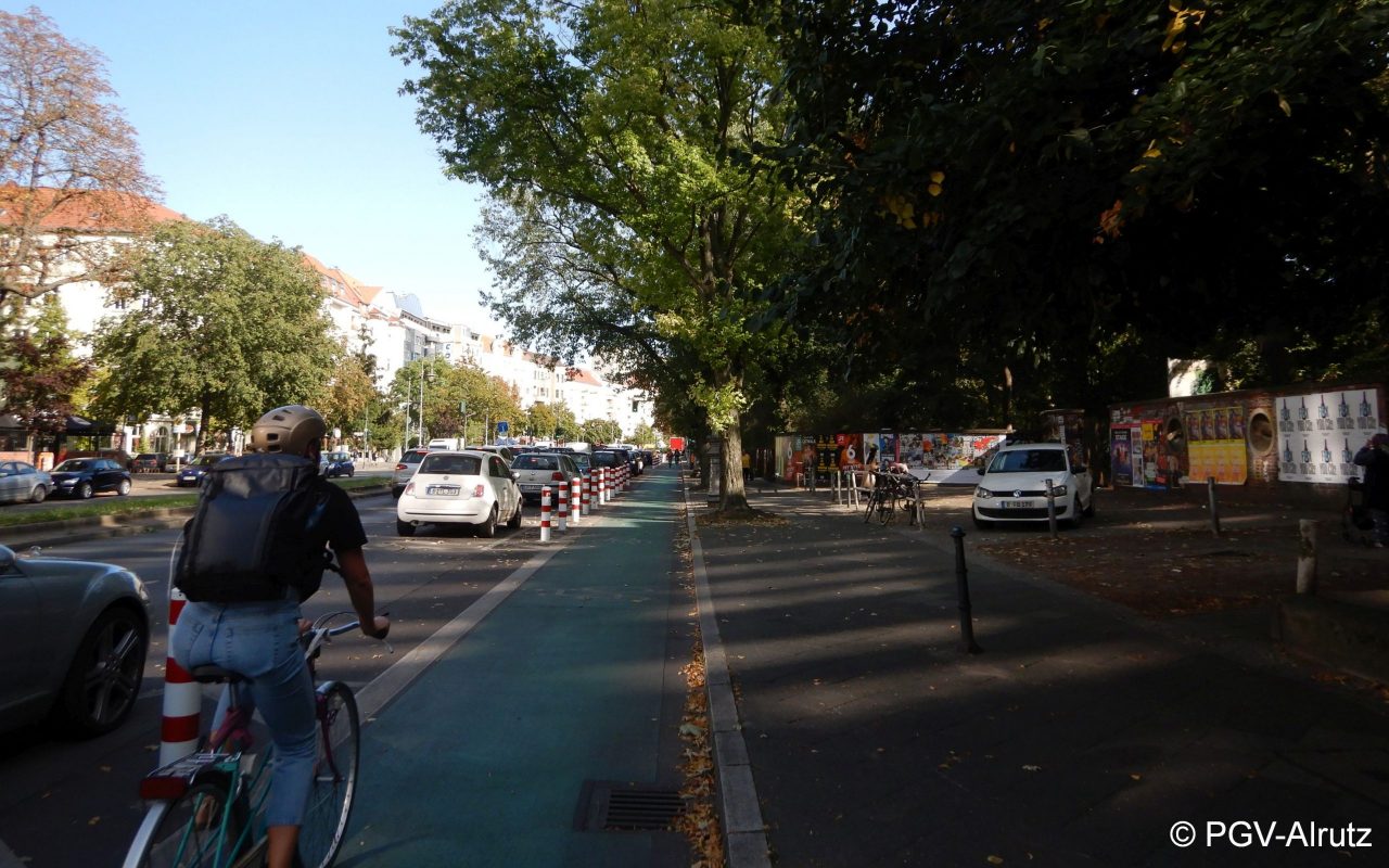 Das Bild zeigt einen geschützten Radfahrstreifen in Berlin. Links ist eine Reihe rot-weiß-gestreifte Poller, die den Radfahrstreifen vom Parkstreifen trennen. Auf dem Radfahrstreifen fährt eine Person Rad- Copyright PGV-Alrutz