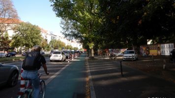 Das Bild zeigt einen geschützten Radfahrstreifen in Berlin. Links ist eine Reihe rot-weiß-gestreifte Poller, die den Radfahrstreifen vom Parkstreifen trennen. Auf dem Radfahrstreifen fährt eine Person Rad- Copyright PGV-Alrutz