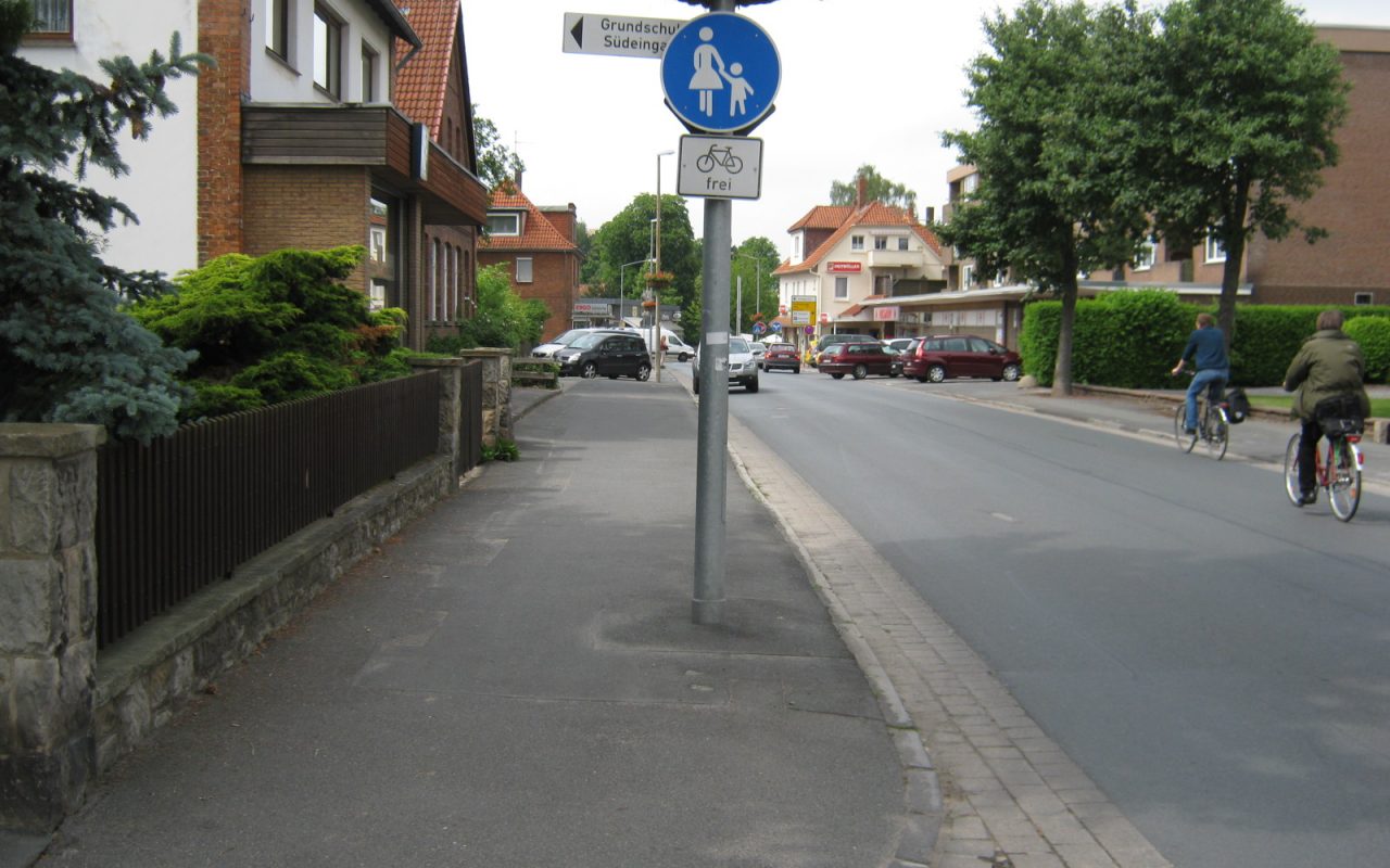 Bild einer Straße mit einem Gehweg (Radverkehr frei) auf der linken Seite und zwei Radfahrenden auf der rechten Fahrbahnseite.