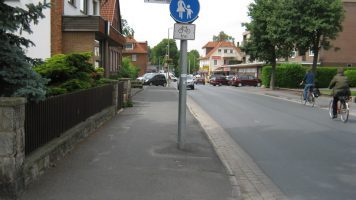 Bild einer Straße mit einem Gehweg (Radverkehr frei) auf der linken Seite und zwei Radfahrenden auf der rechten Fahrbahnseite.
